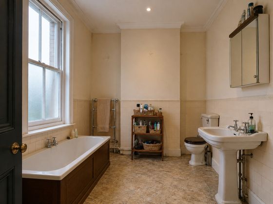 Outdated larger Victorian British bathroom with a tall sash window, built-in bath, pedestal basin, toilet, cream half-height tiles and tired flooring before renovation.